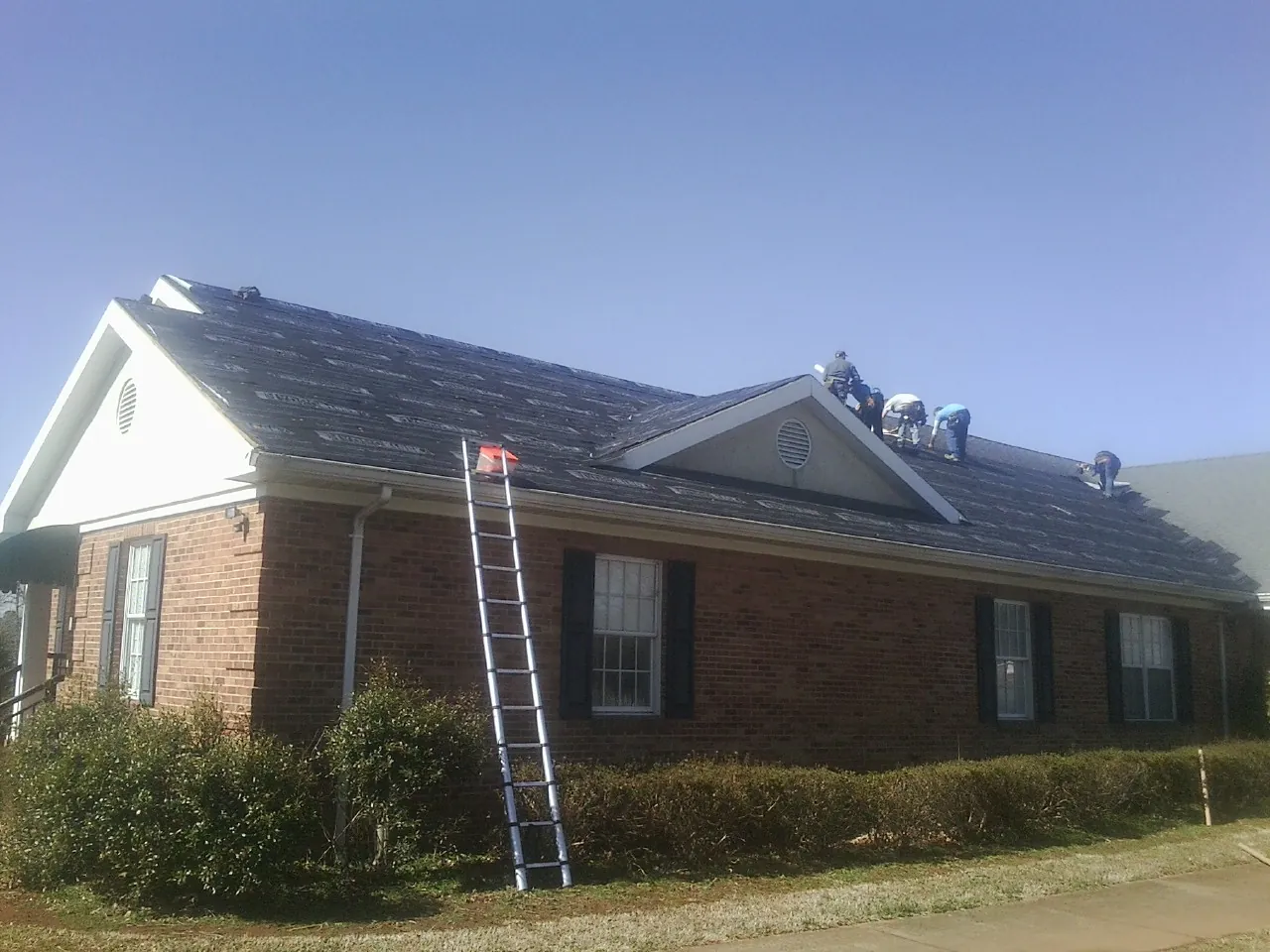 Crew tearing shingles off a brick ranch roof with a ladder set — Emergency Roof Repair work by our crew in Gateway